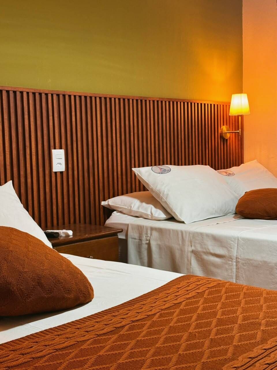 Una habitación de hotel con cabecera de madera, dos camas con sábanas blancas, una mesita de noche y una lámpara de pared.