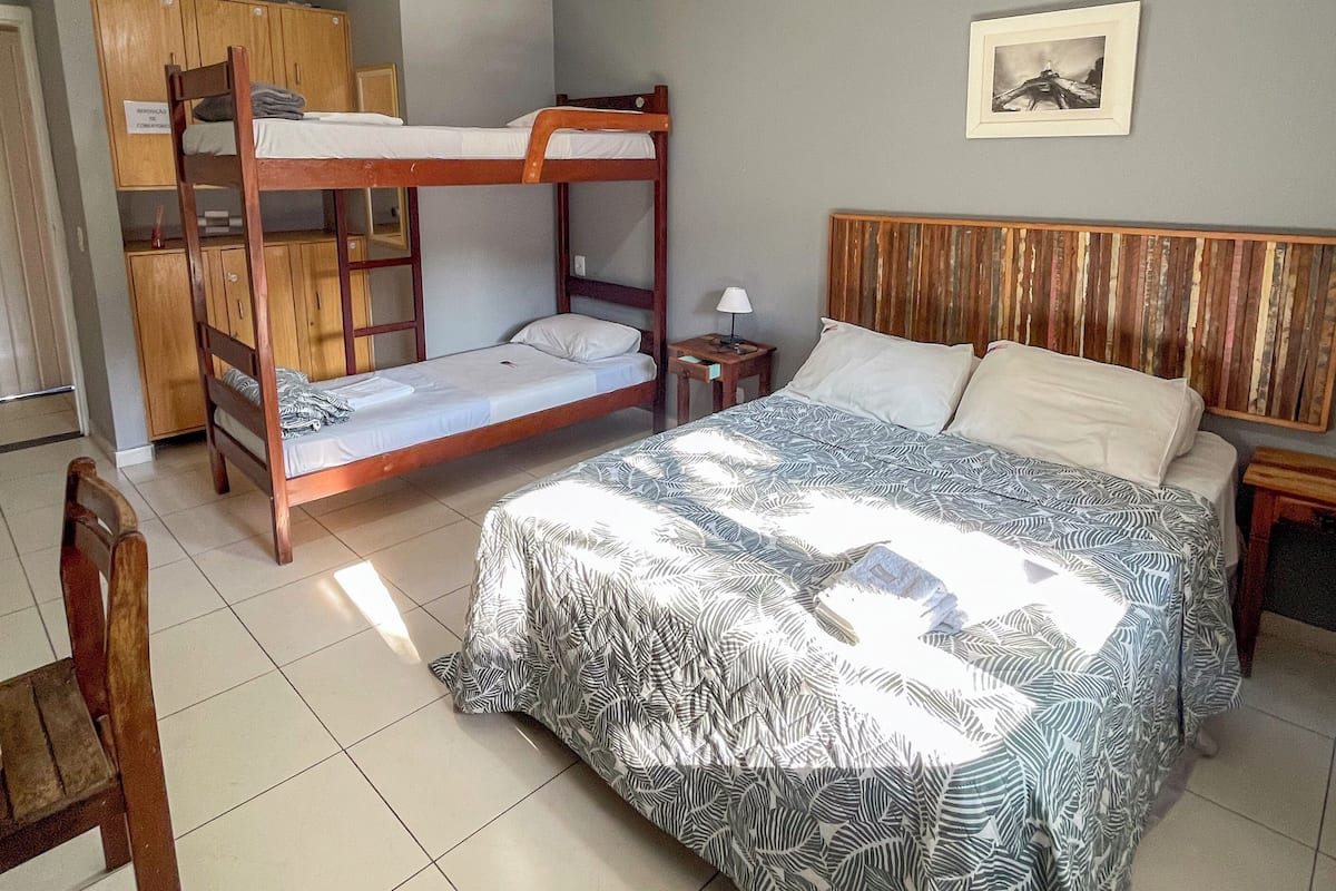 A dorm-style room with bunk beds, a single bed, a wooden wardrobe, a framed picture on the wall, and a tiled floor.
