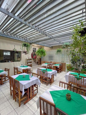 Un restaurante con mesas cubiertas de manteles blancos y caminos de mesa verdes, sillas de madera y plantas colgantes.