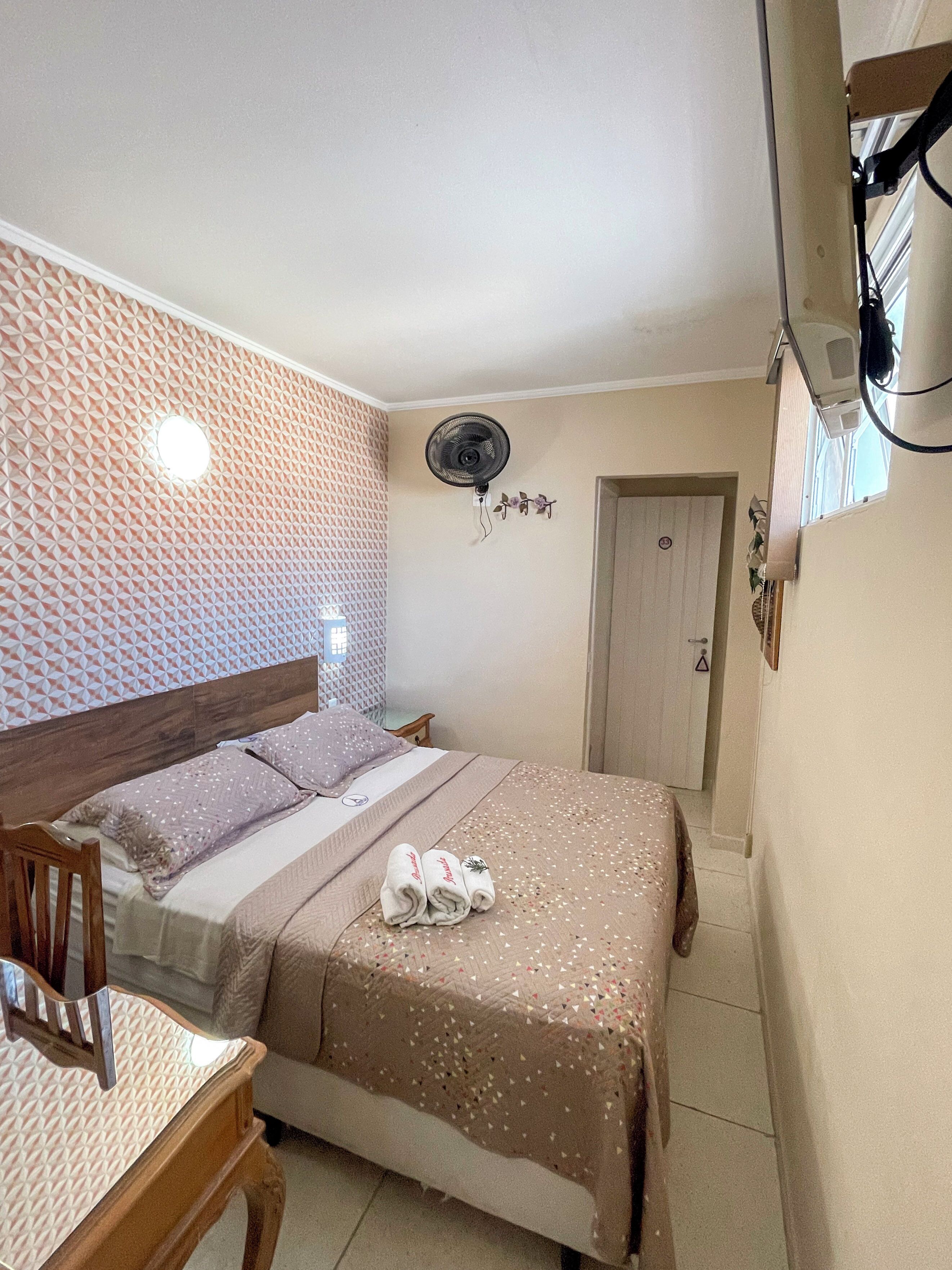 A neatly made bed with a patterned bedspread, a wall-mounted fan, and a door with a security lock.