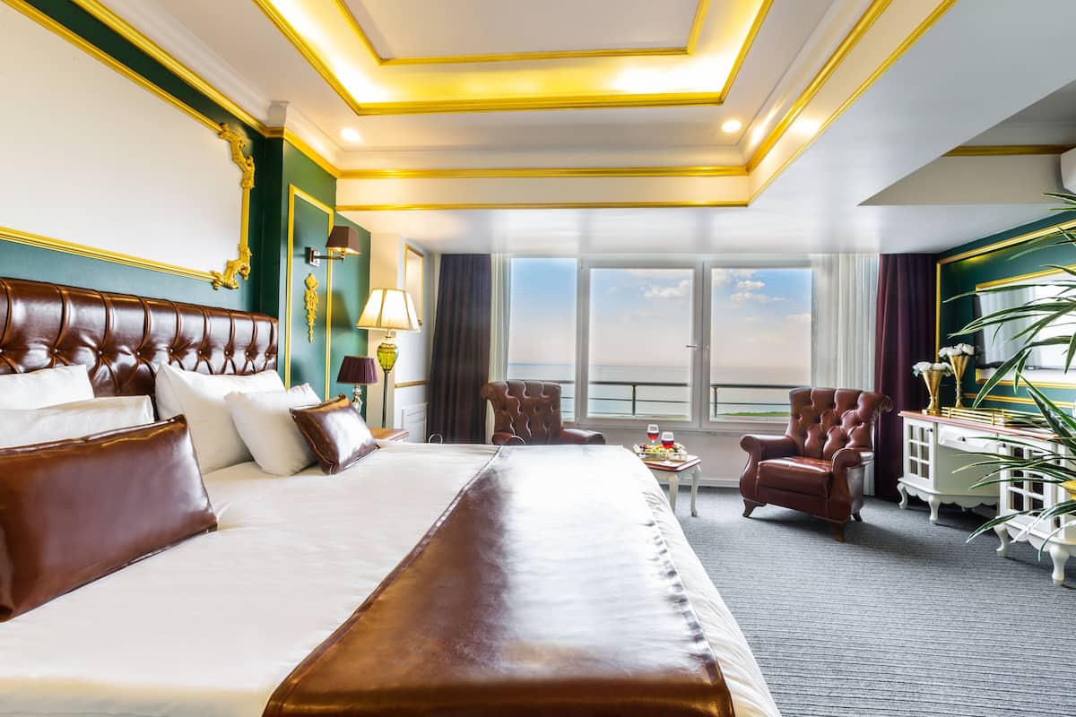 A large bed with a headboard, a sitting chair, a small table with a lamp, and a view of the sky through large windows.