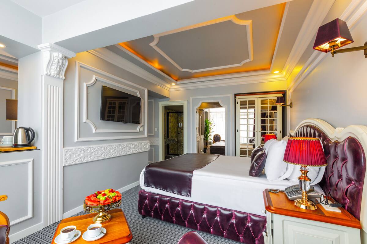 A hotel room with a large bed, a bedside table with a lamp, a small table with a fruit bowl and cups, and a mirror on the wall.