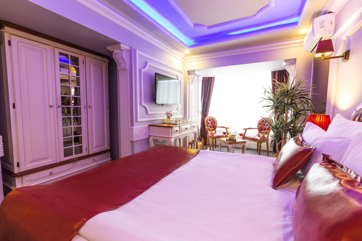 A hotel room with a large bed, a wardrobe, a desk with a chair, a television, and a view of the outside.