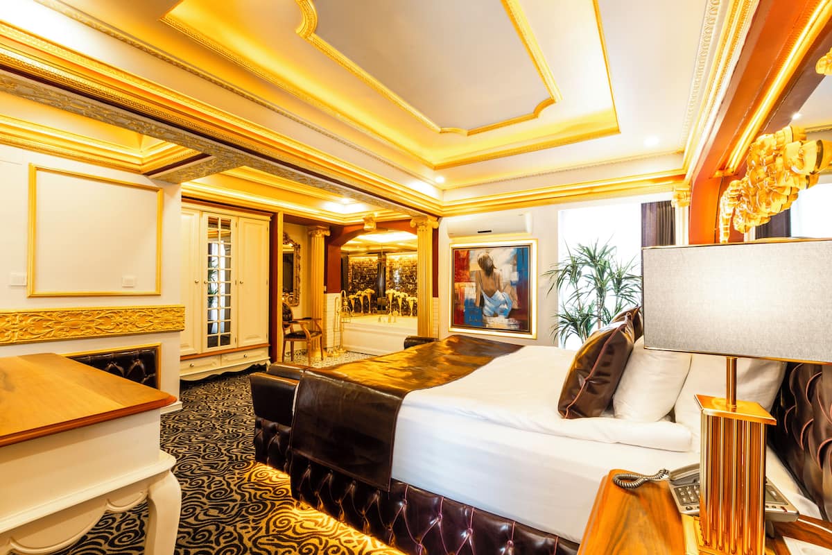 A luxurious hotel room with a large bed, a sitting area with a sofa, a dining table, and a view of the interior.