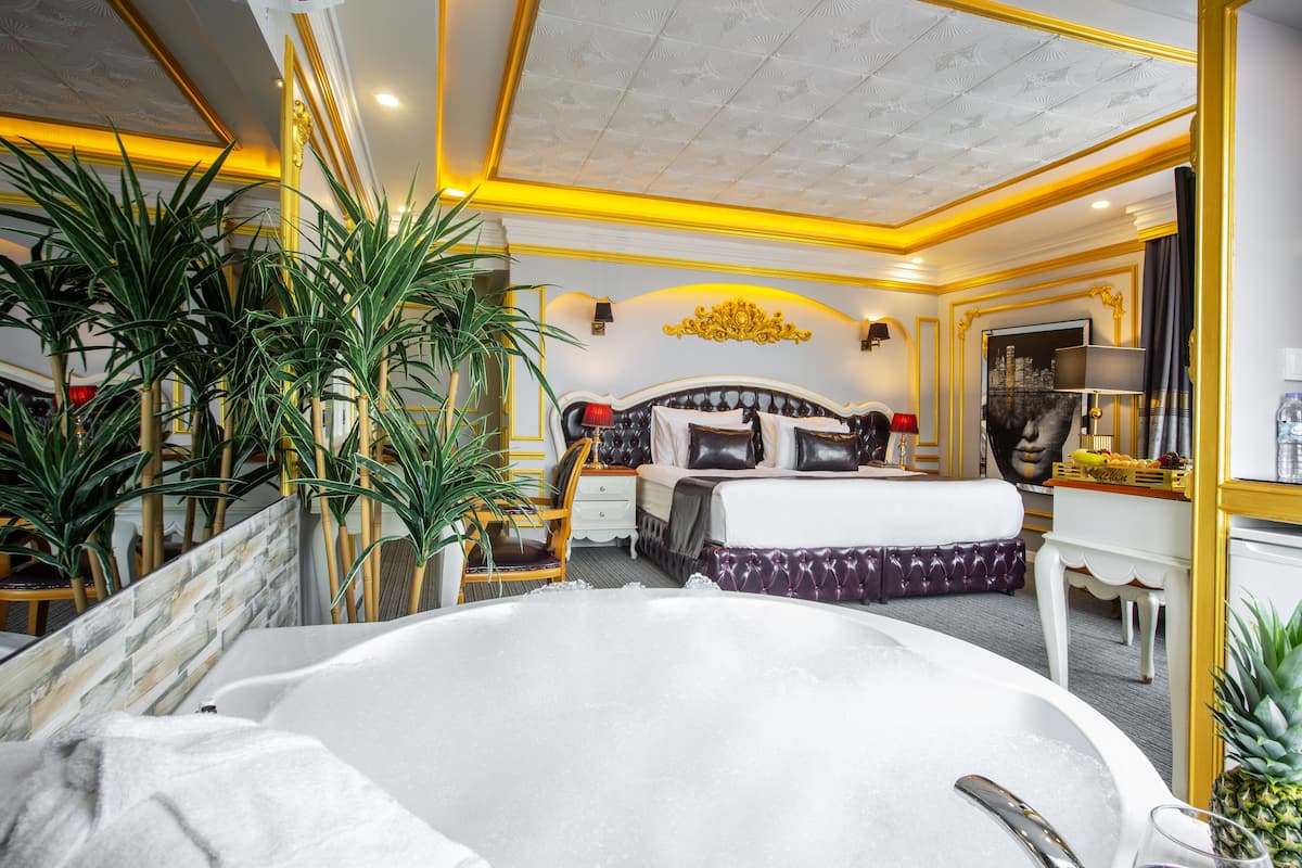 A luxurious bedroom with a large bed, a sitting area with a chair and table, a bathtub, and a dining area with a fruit basket.