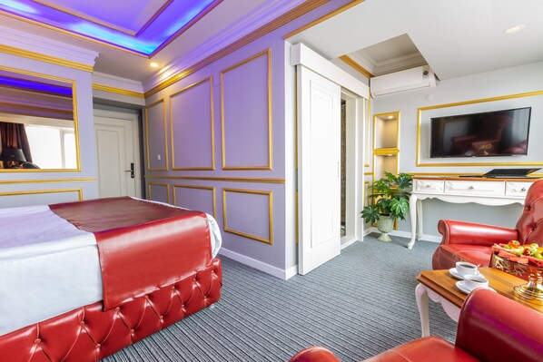 Habitación de hotel con cama de cuero rojo, televisor, escritorio con ordenador portátil y zona de comedor con una silla y mesa roja.