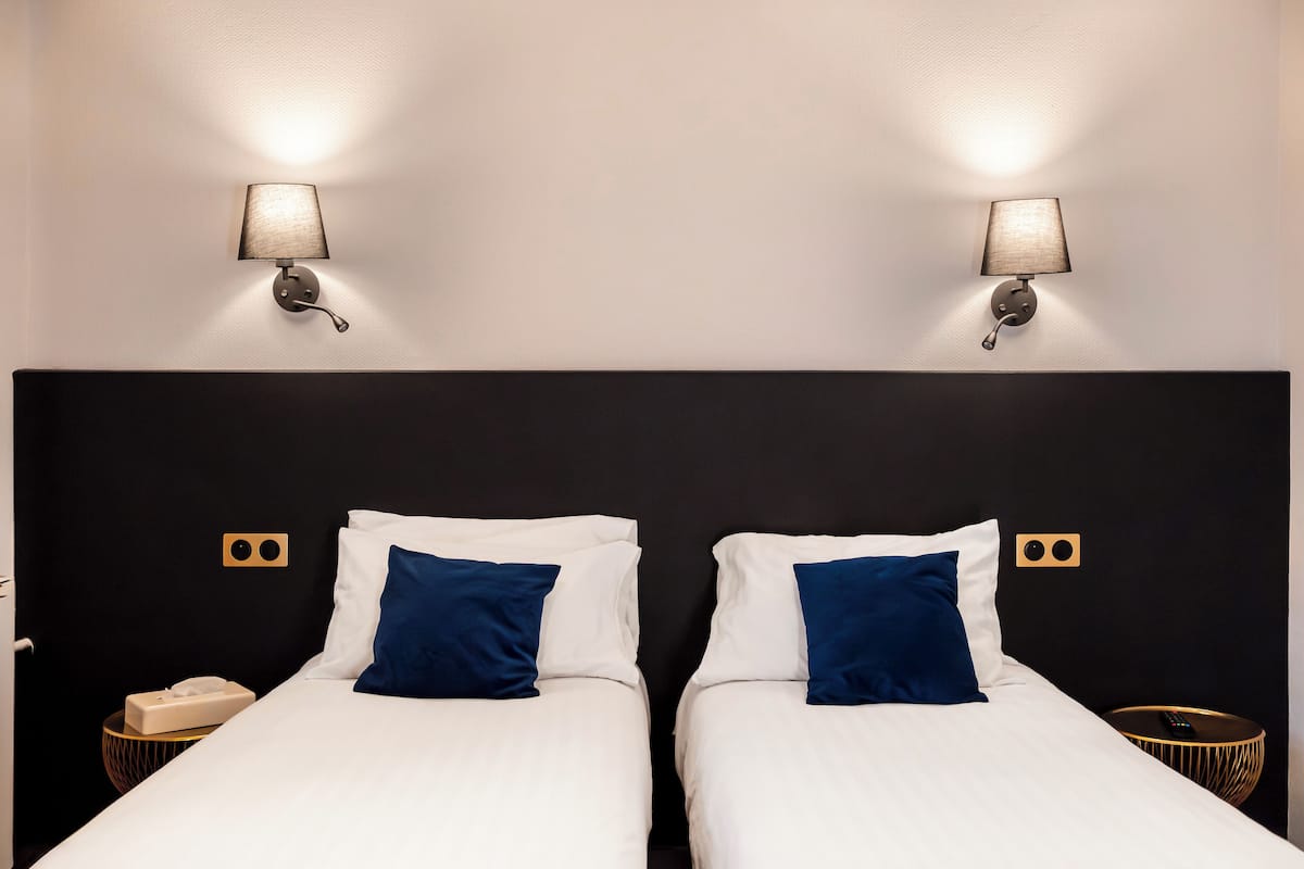 Une chambre d’hôtel avec deux lits, chacun agrémenté de draps blancs et d’un seul oreiller bleu. Deux lampes murales sont fixées de part et d’autre des lits.