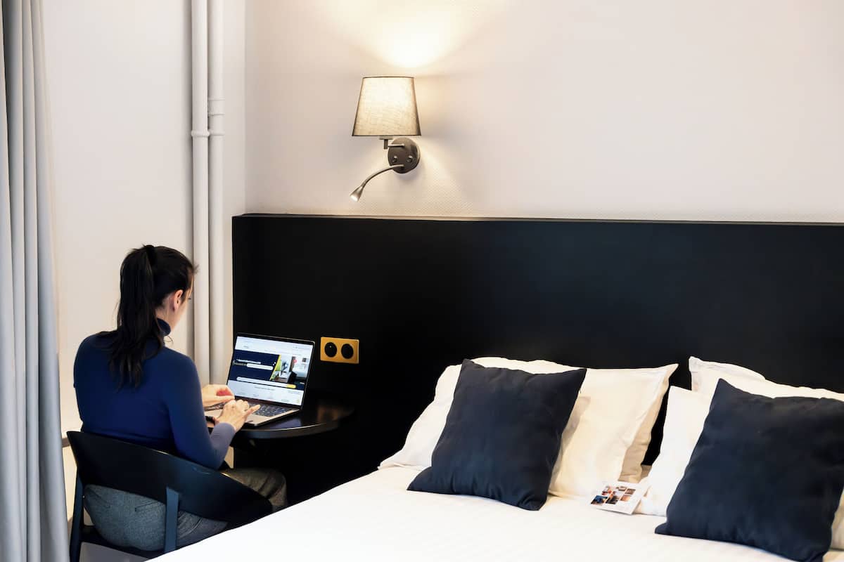 Une chambre d’hôtel équipée d’un lit, d’un bureau avec un ordinateur portable, d’une lampe et d’un haut-parleur fixé au mur.