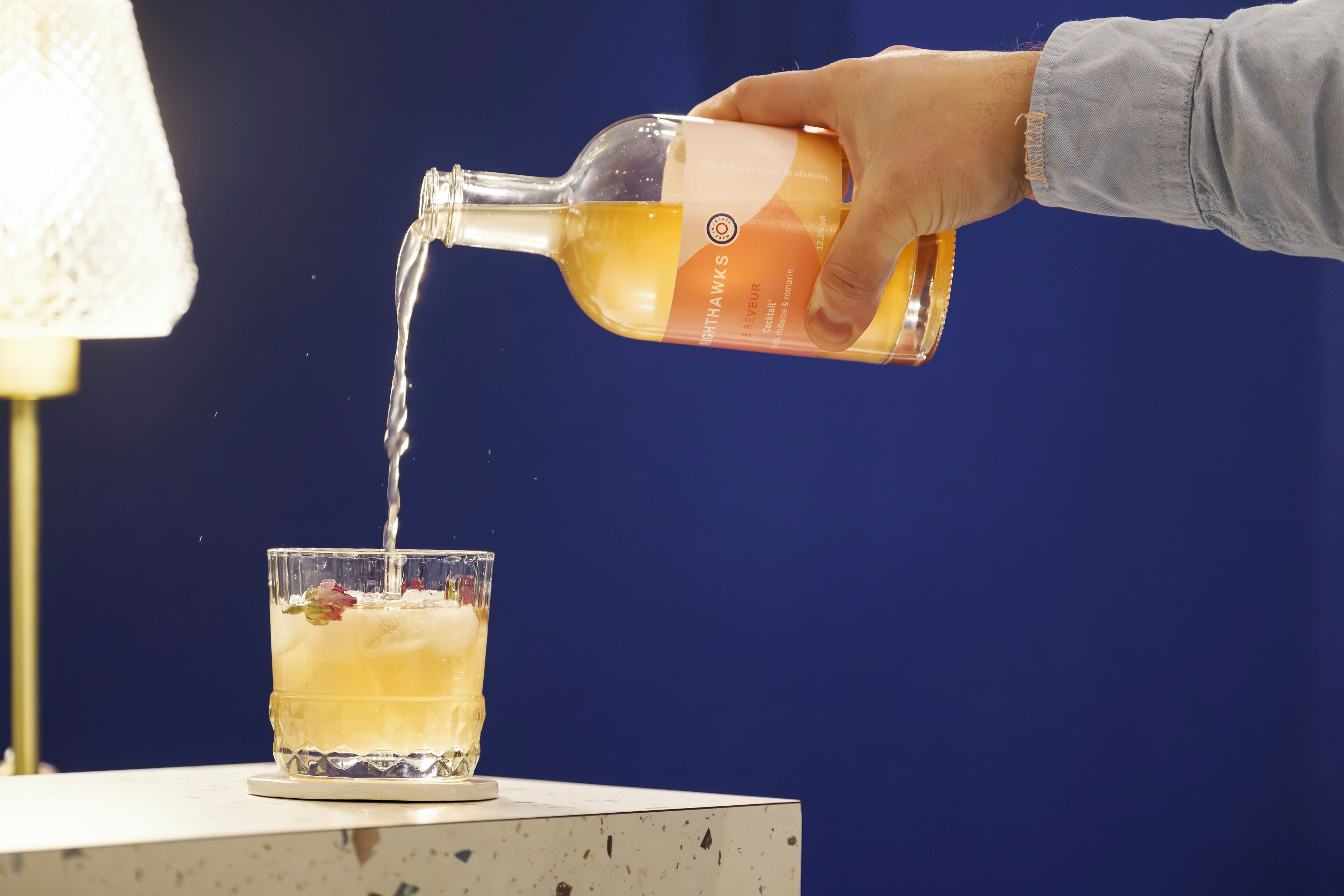 Una mano vertiendo un líquido transparente de una botella en un vaso con hielo y una flor como adorno.