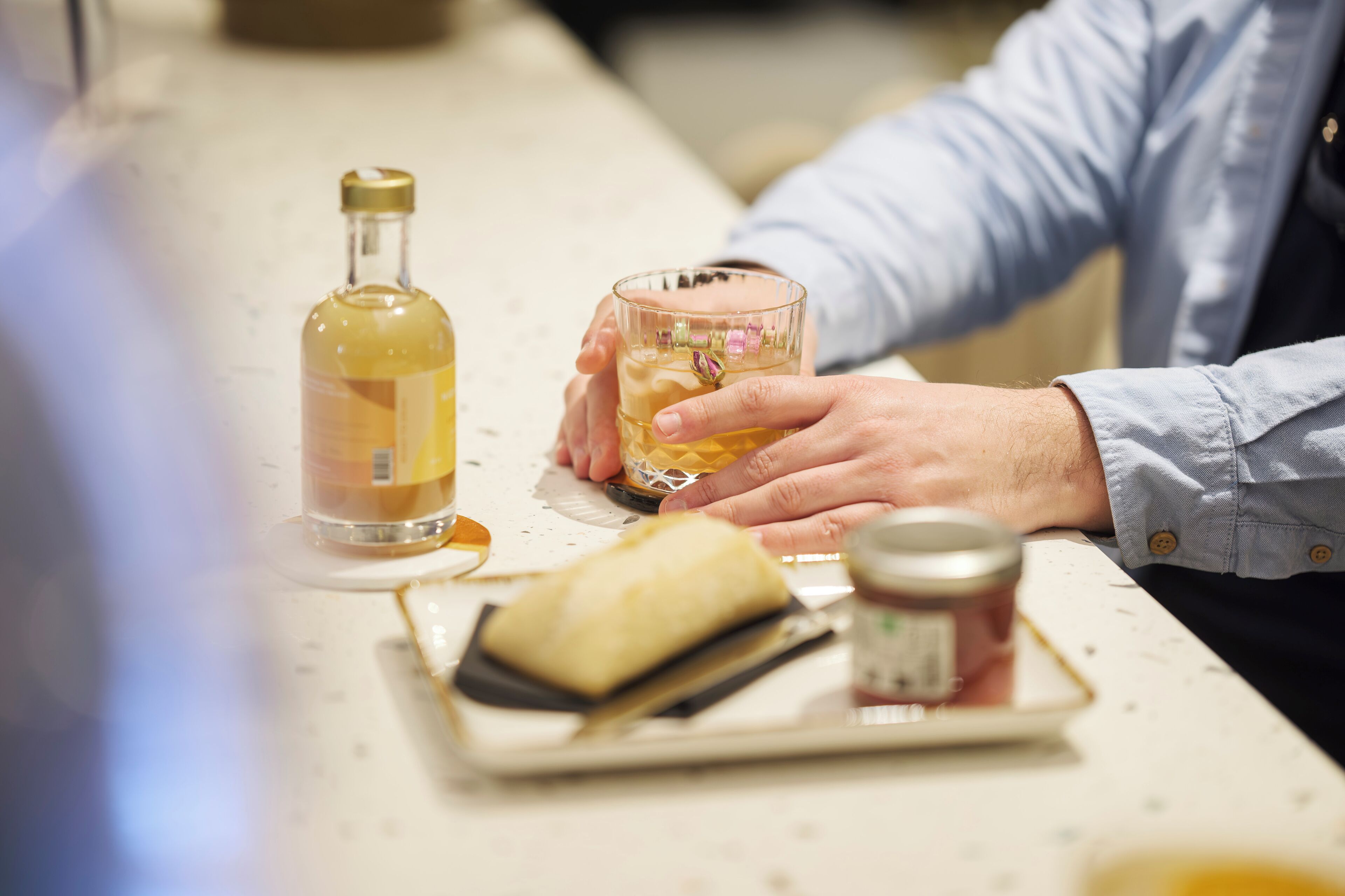Una persona sosteniendo un vaso con una bebida, una botella y un pequeño plato con un pastel sobre una mesa.