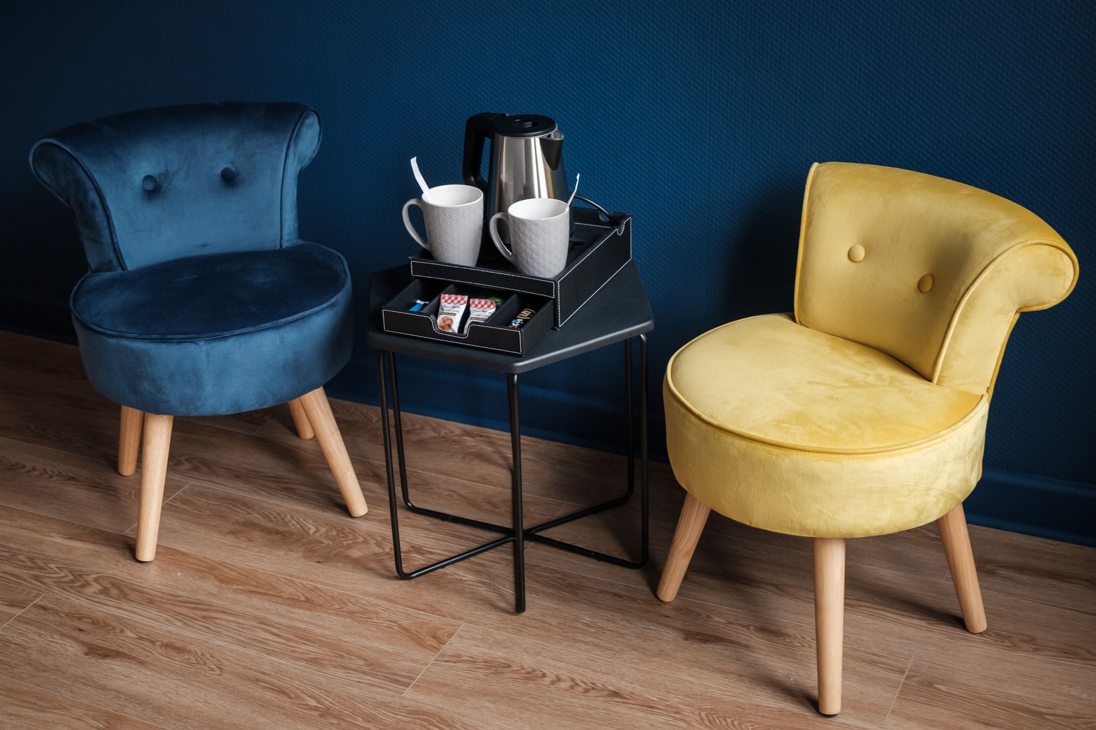 Two chairs, one blue and one yellow, with a small table between them holding a coffee pot, cups, and a box of tissues.