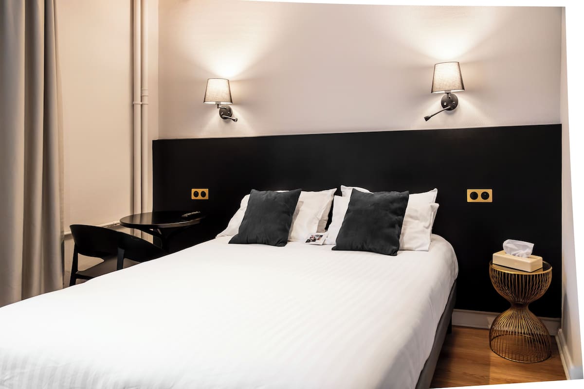 Un lit bien fait, avec une literie blanche et grise, deux lampes fixées au mur, une petite table ronde avec une chaise, et une table de chevet avec une boîte à mouchoirs.