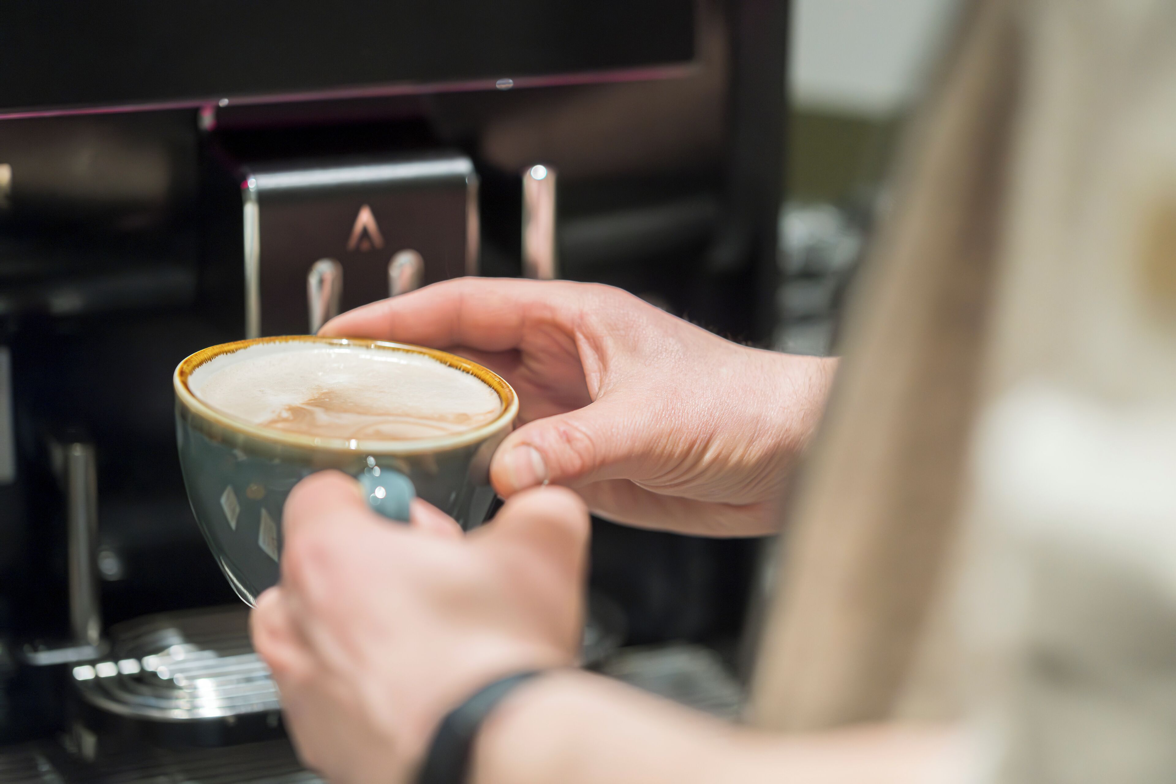 A person holding a cup of coffee with a frothy top, likely from an espresso machine.