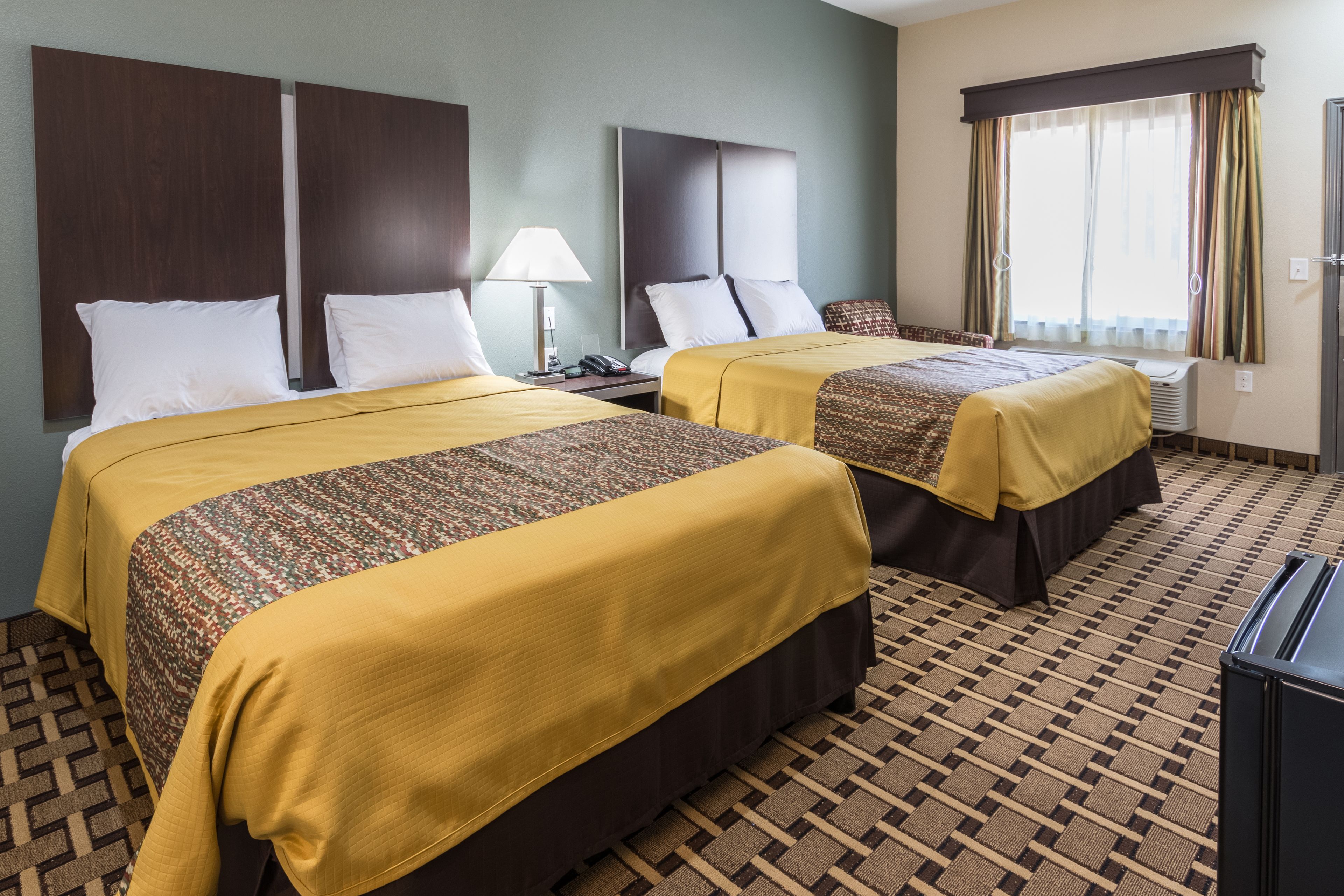A hotel room with two beds, a patterned carpet, a window with curtains, and a desk with a lamp.