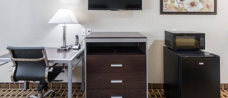 A hotel room with a desk, chair, lamp, TV, microwave, and a small refrigerator.