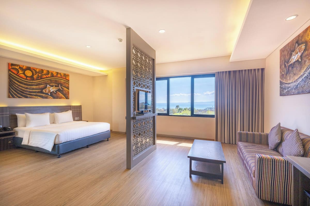 A modern hotel room with a large bed, a sofa, a small table, and a view of the sea.