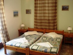 A double bed with a patterned bedspread, two bedside tables with lamps, a window with curtains, and a wall with two framed pictures. - Pension Lagoudera (Naxos)