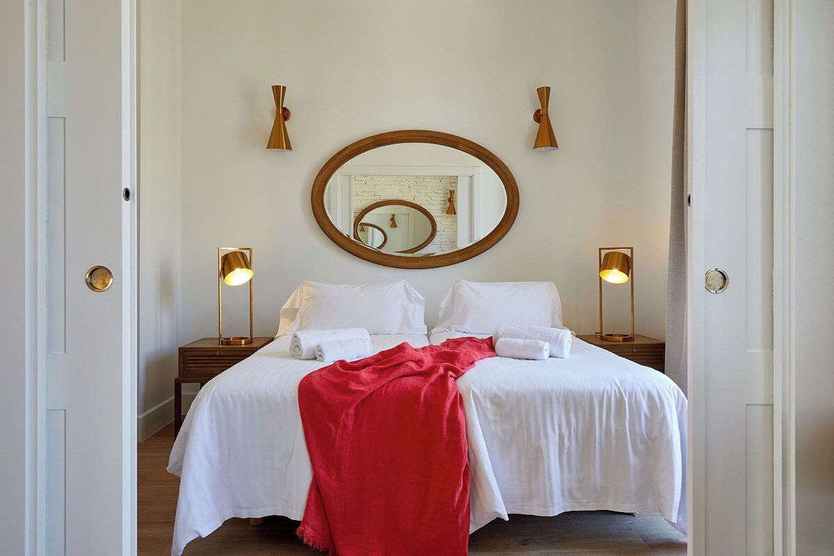 Une chambre d’hôtel avec un grand lit, deux tables de chevet, un miroir rond et deux lampes fixées au mur.