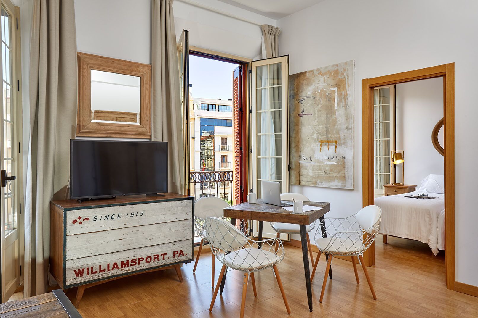 Hotelkamer met een houten dressoir, een televisie, een eettafel met stoelen, een bed en een groot raam met uitzicht op gebouwen.