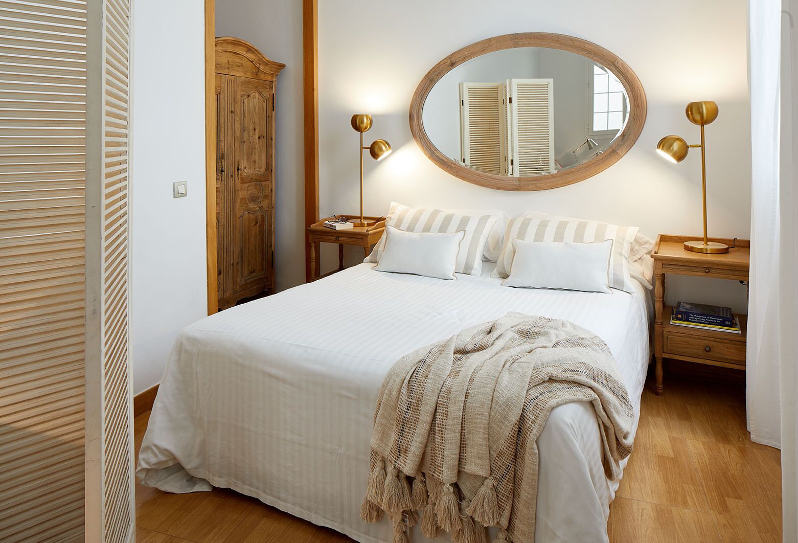 A neatly arranged bedroom with a large bed, a wooden wardrobe, a round mirror, and bedside lamps.