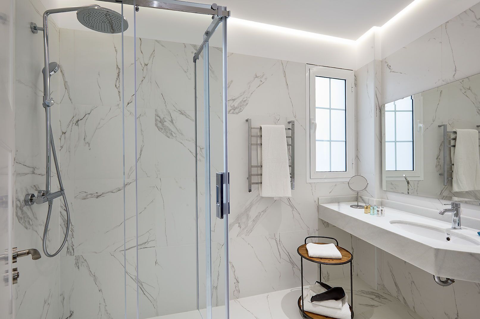 A modern bathroom with a glass-enclosed shower, a marble-tiled wall, a large mirror, and a vanity with a sink and towel rack.