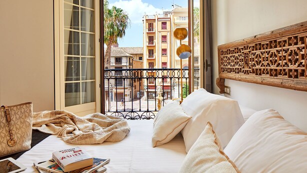 Habitación de hotel con una cama, vista a la calle, cabecero de madera y ventana con vistas a edificios y palmeras.