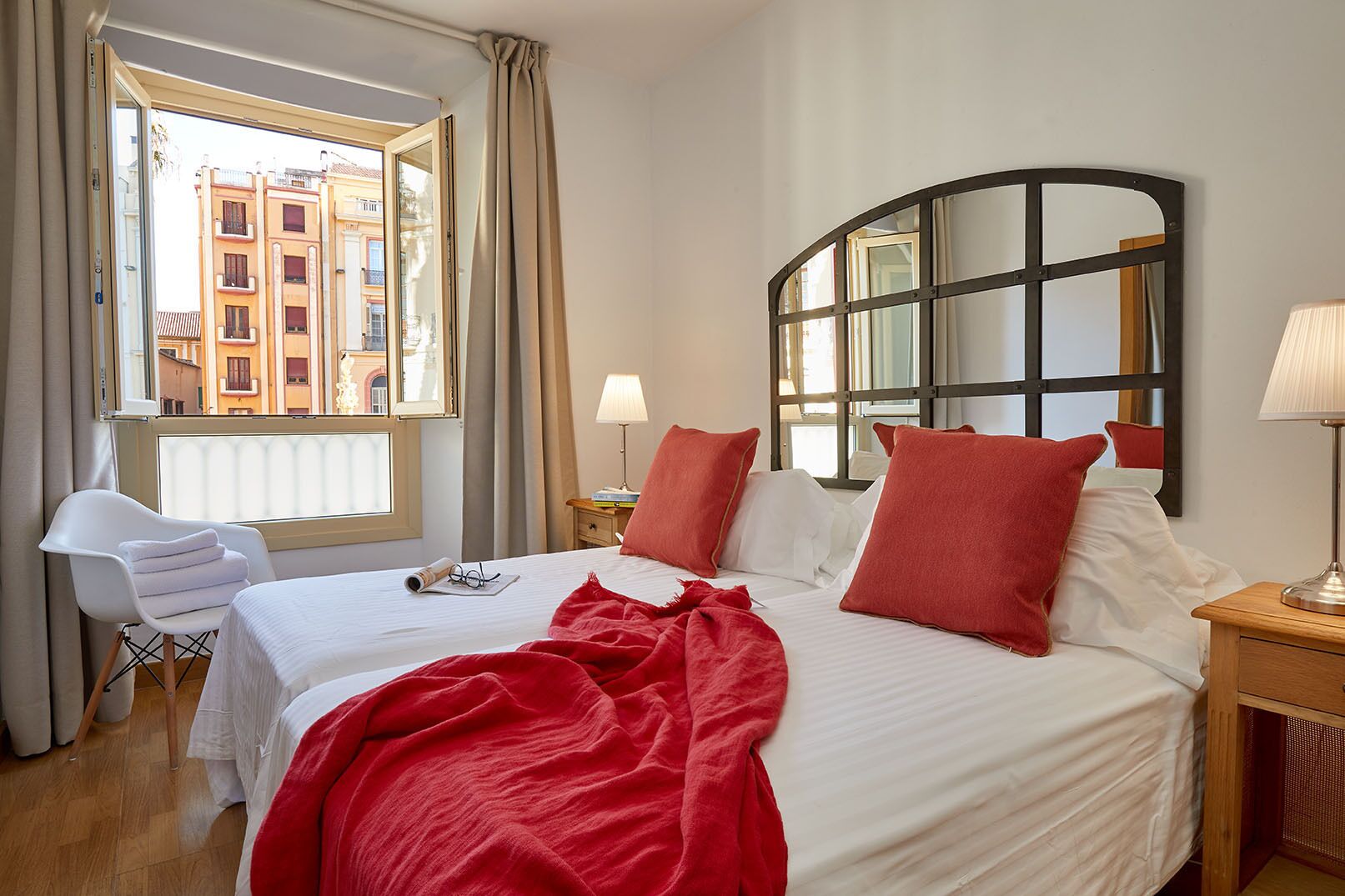 A hotel room with a bed, a chair, a window with curtains, a lamp, and a view of buildings outside.