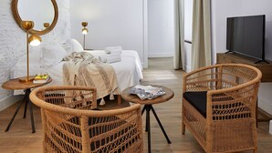Habitación de hotel con una cama, dos sillas de mimbre, una mesa redonda, un espejo y un televisor.