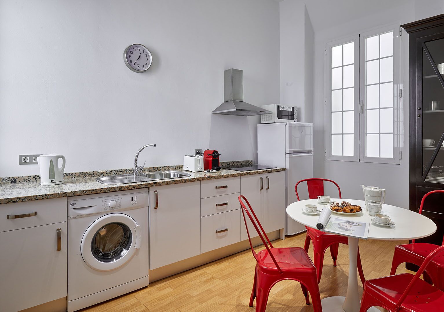 Eine kompakte Küche mit Waschmaschine, Mikrowelle und Spüle. Ein Esstisch mit roten Stühlen und ein Fenster mit weißen Rahmen.