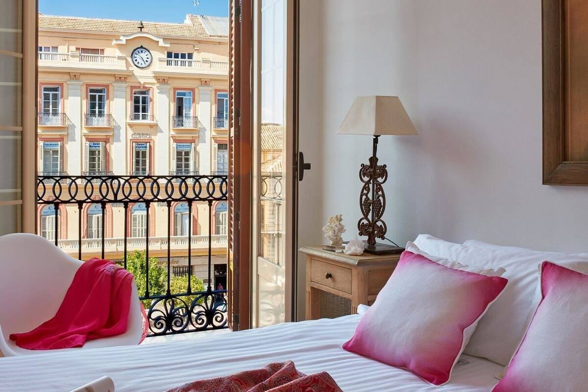 Habitación de hotel con cama, una silla, mesita de noche, lámpara y vistas a un edificio con balcones.