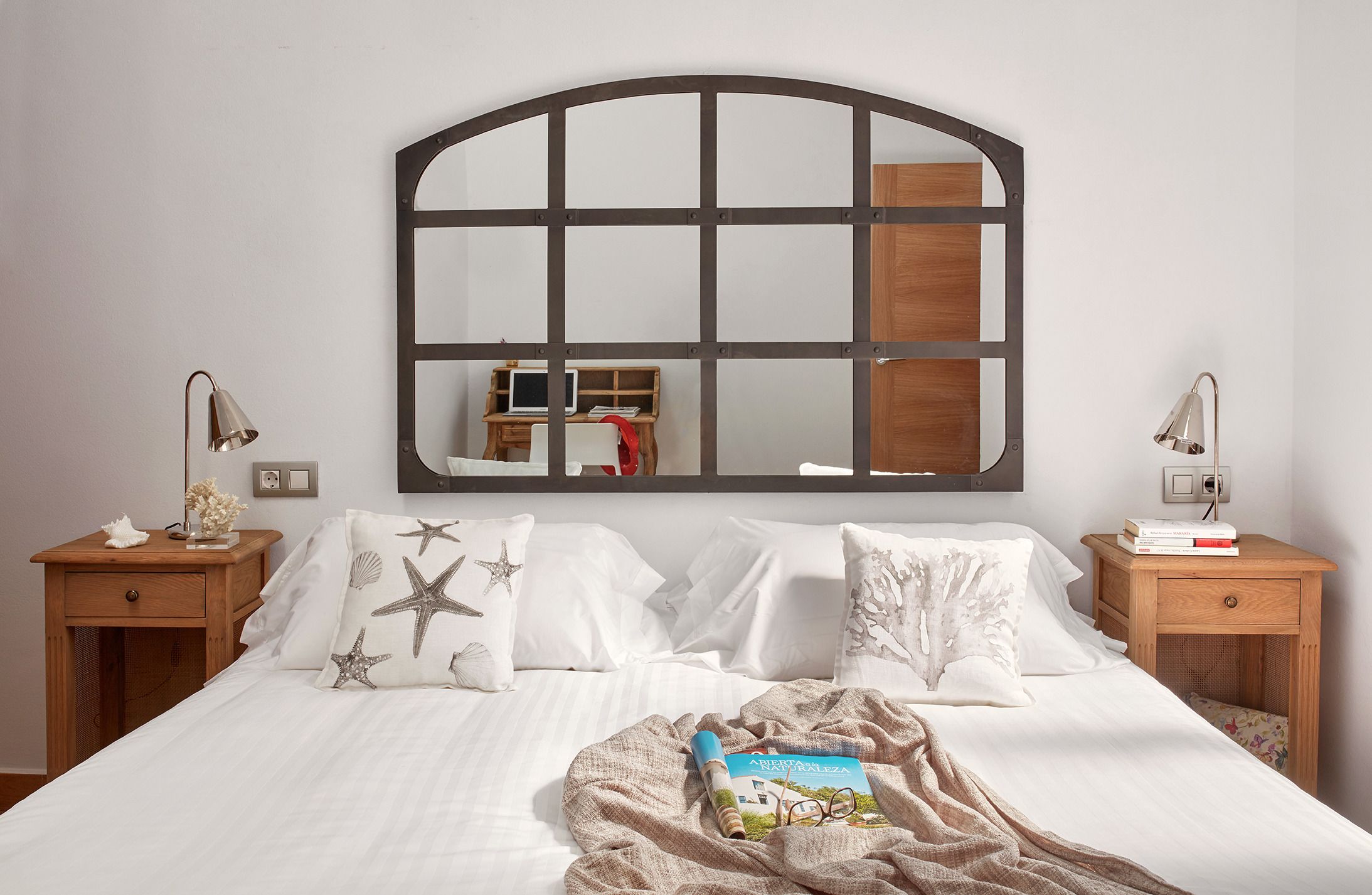 Uma cama bem arrumada com um espelho grande acima, duas mesas de cabeceira com abajures e um painel de madeira na cabeceira.