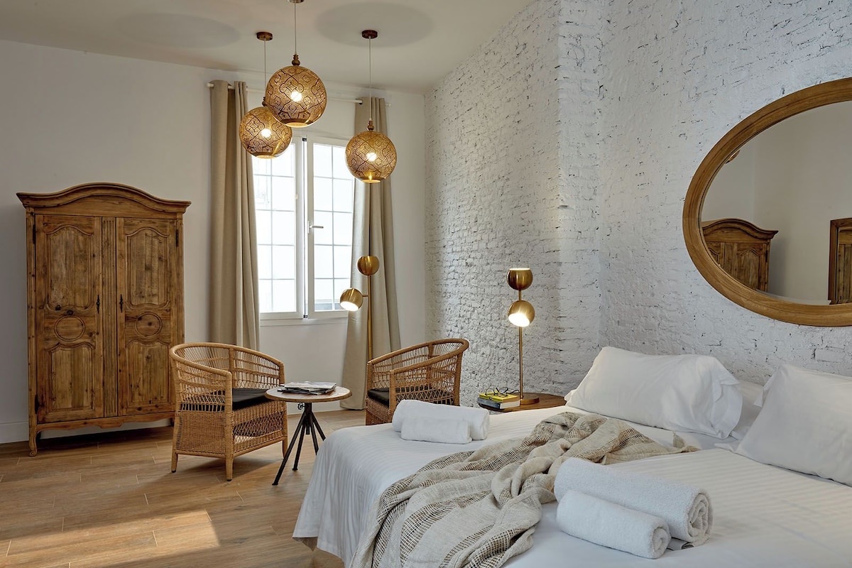Une chambre avec deux lits, une armoire en bois, des chaises en osier, un miroir rond et un mur en briques.