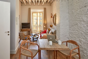 A cozy living room with a wooden table, two wicker chairs, a sofa, a TV, and a balcony with a view.