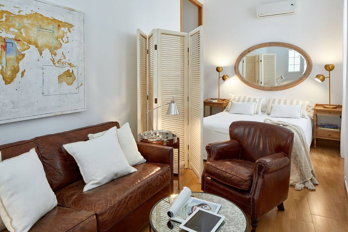Habitación de hotel con sofá de cuero marrón, una mesa redonda con una tablet y un libro, suelo de madera, cama con ropa de cama blanca, lámpara de noche, espejo redondo y un mapa en la pared.