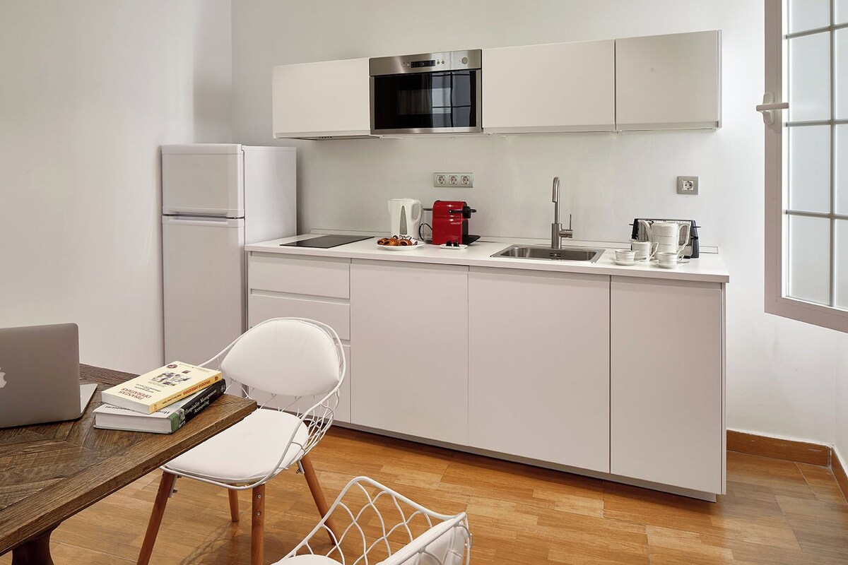 Une cuisine moderne avec des meubles blancs, un four en acier inoxydable, une machine à café et un ordinateur portable posés sur une table en bois.