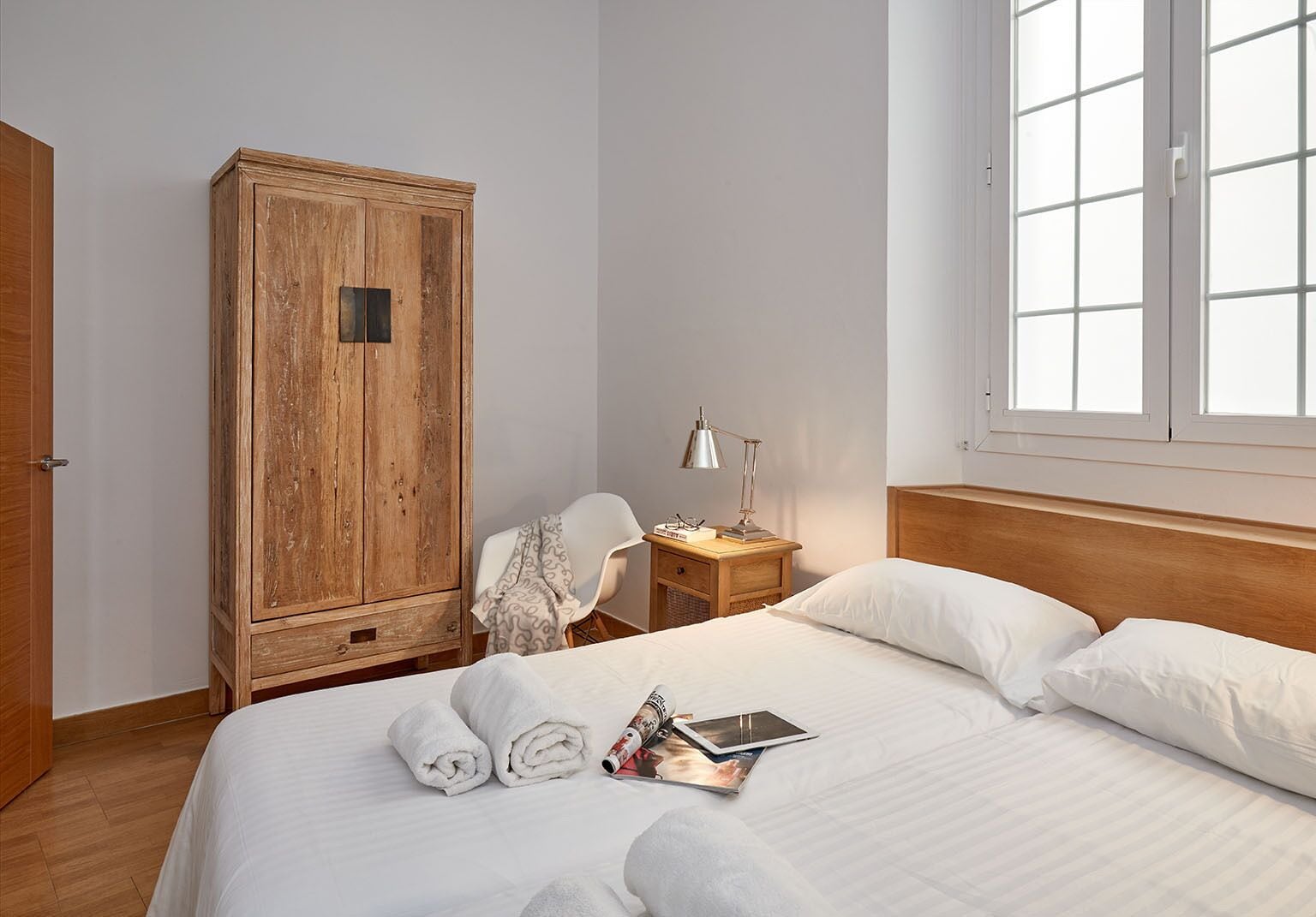 Una cama bien hecha con ropa blanca, un armario de madera, una mesita de noche con una lámpara y una ventana con vistas.