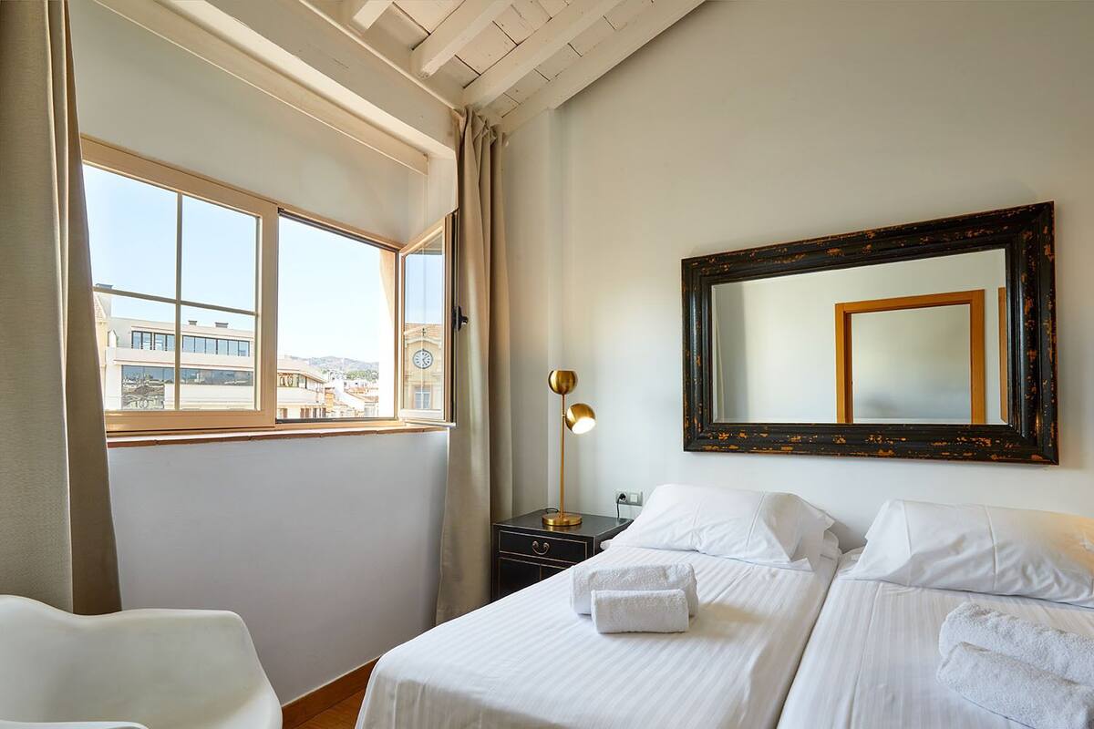 Habitación de hotel con dos camas, un ventanal con vistas a edificios, un escritorio con lámpara y un espejo en la pared.