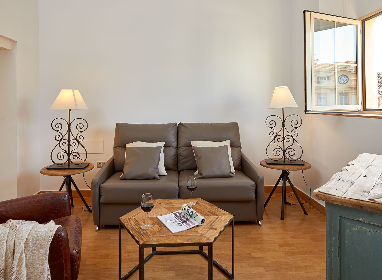Un salon comprenant un canapé, deux tables d’appoint avec des lampes, une table basse avec un verre de vin et un magazine, et une vue sur un immeuble par la fenêtre.