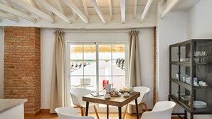 Un comedor con pared de ladrillo, una mesa de madera, sillas blancas y un aparador de vidrio con vajilla.
