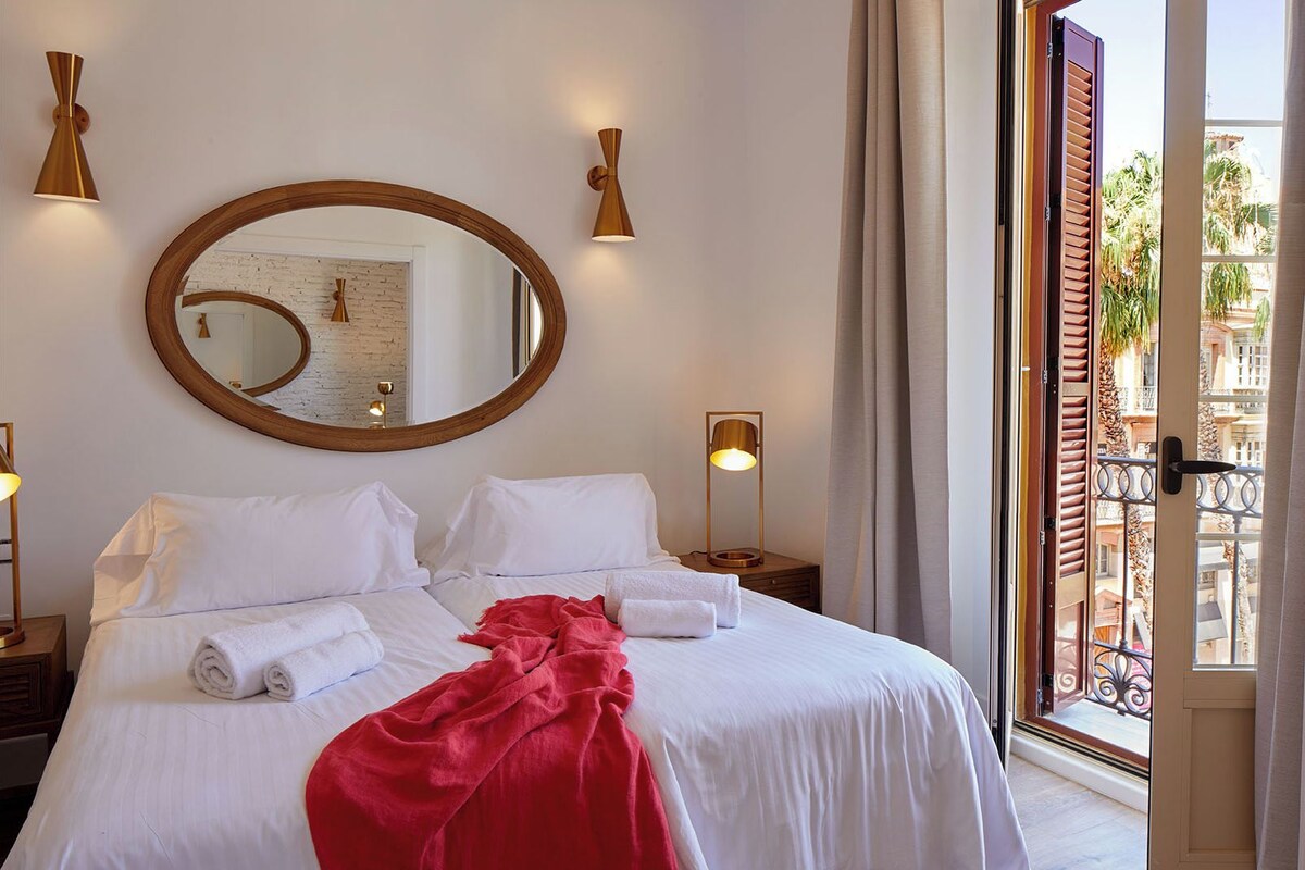 A hotel room with a large bed, a round mirror, two bedside lamps, and a view of the outside balcony.