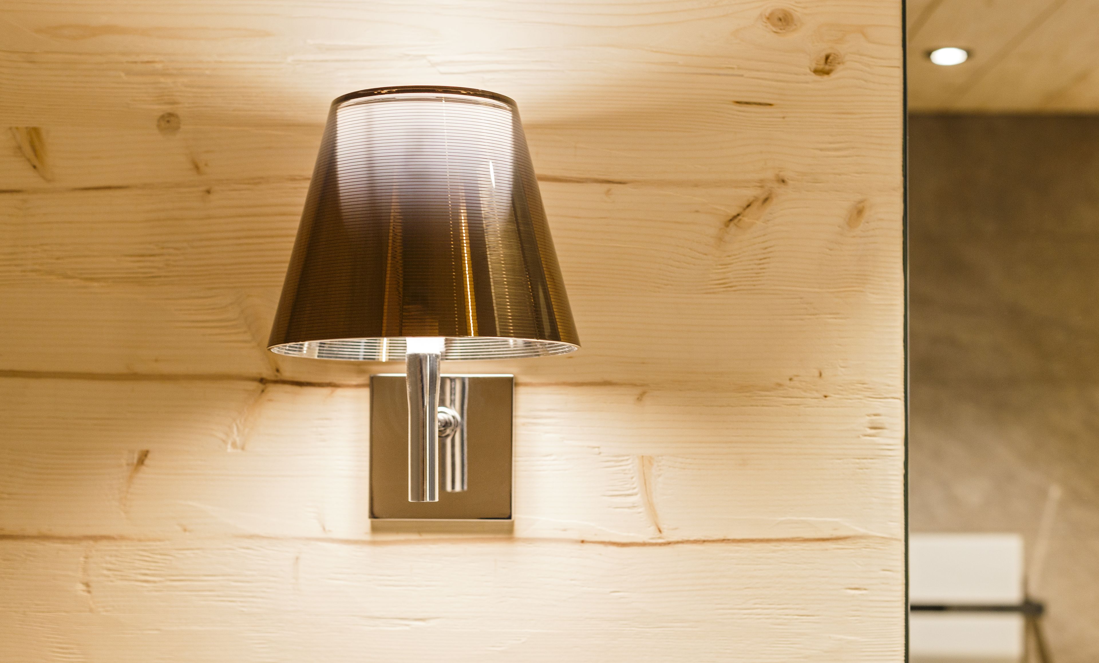 A metallic wall-mounted light fixture with a conical shade against a wooden wall.