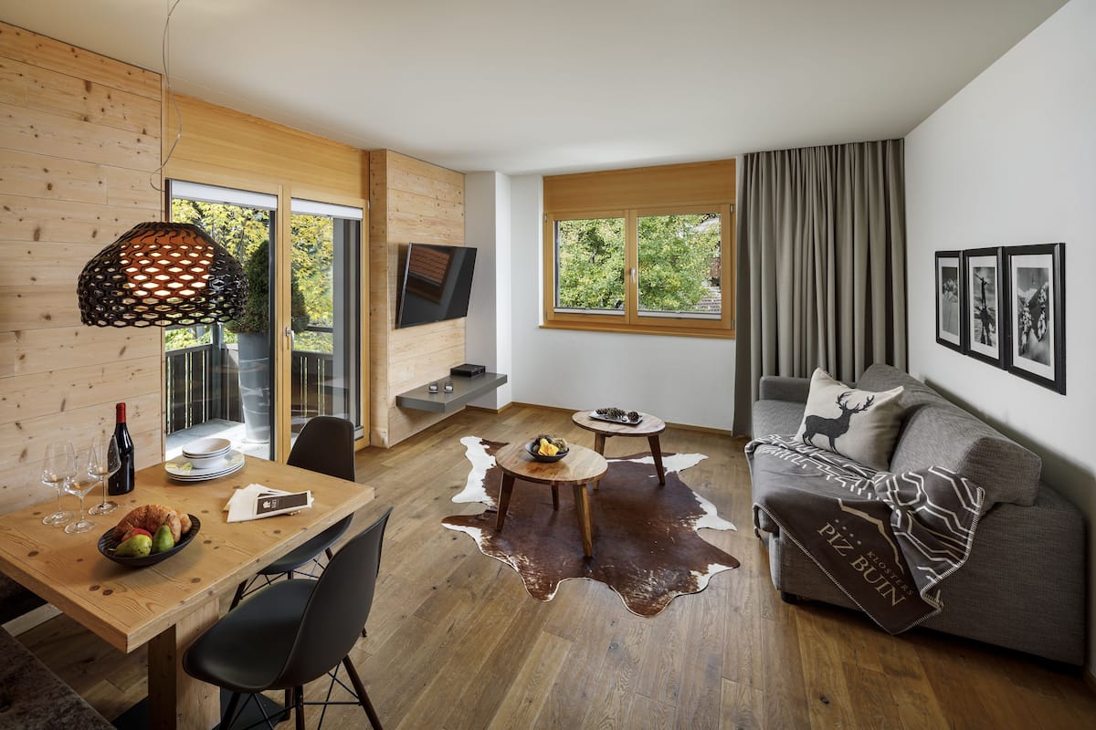 Ein modernes Wohnzimmer mit einem Holzesstisch, einem Sofa und einem Kuhfellteppich, sowie einem großen Fenster mit Vorhängen.