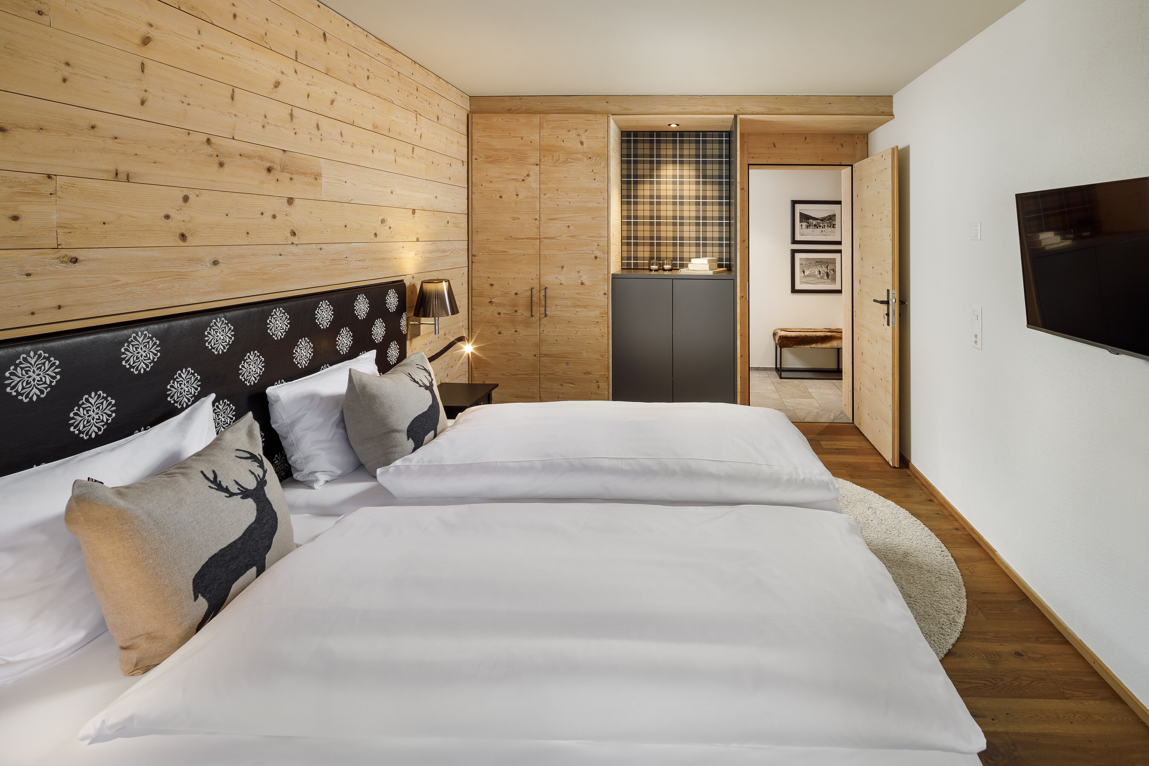 A bedroom with a wooden headboard, two beds with white bedding, a bedside table with a lamp, and a television on the wall.
