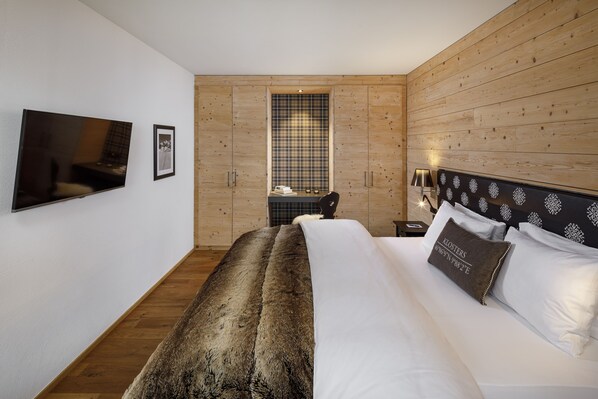 A bedroom with a large bed, a TV mounted on the wall, a window with a plaid curtain, and wooden paneling on the walls.