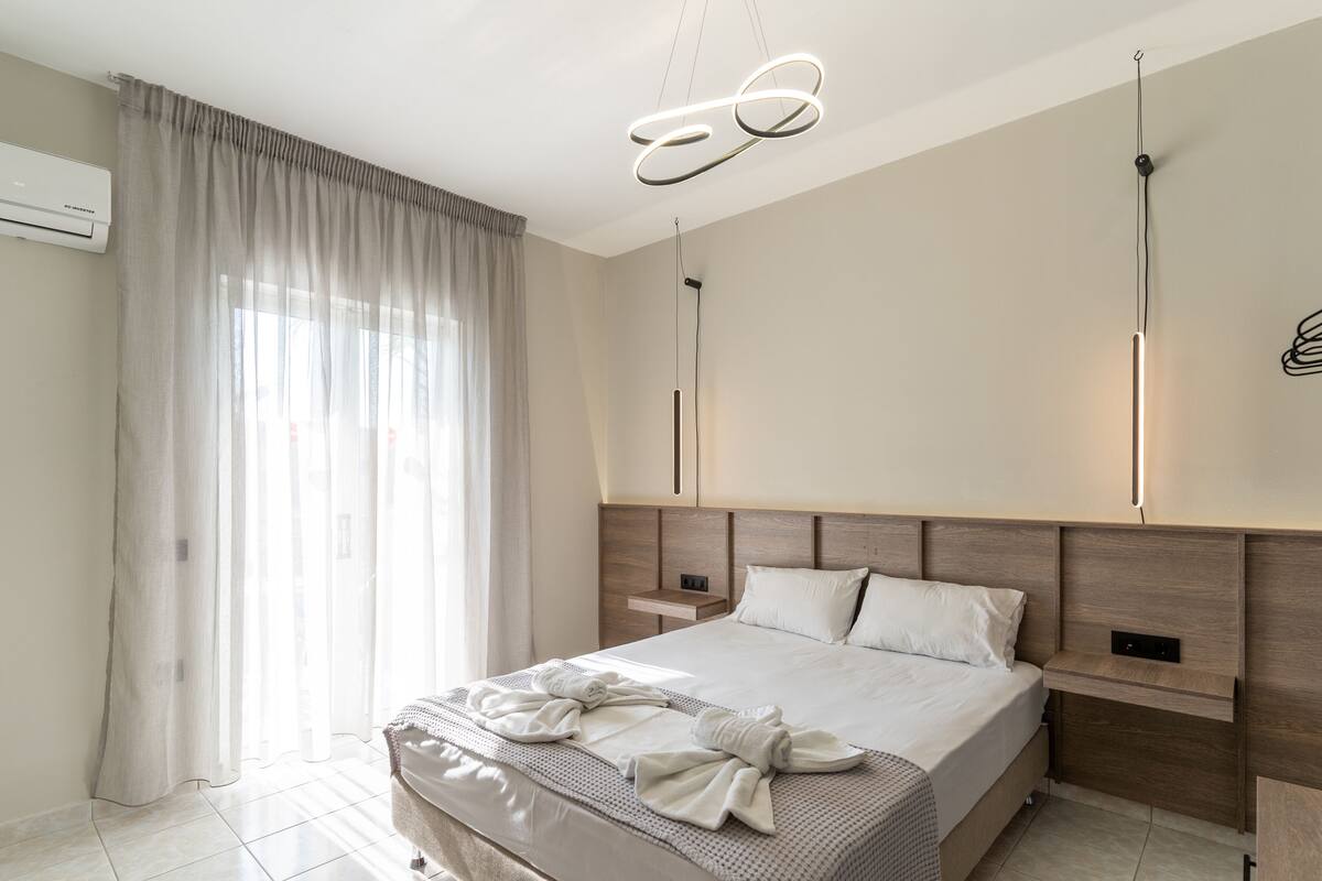A modern hotel room with a large bed, a bedside table, a nightstand, a wall-mounted light fixture, and a window with sheer curtains.