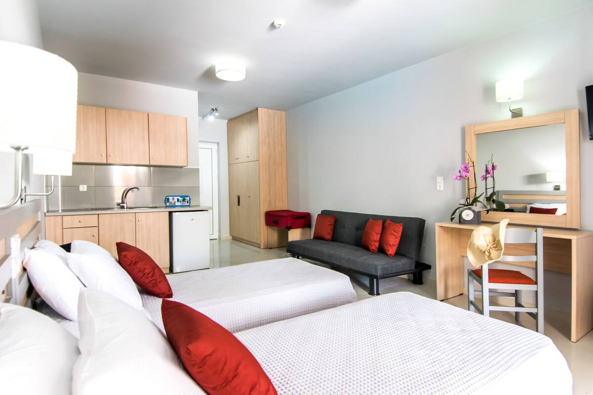 A modern hotel room with a kitchenette, two beds with red and white bedding, a gray sofa, a wooden desk with a chair, and a large mirror.