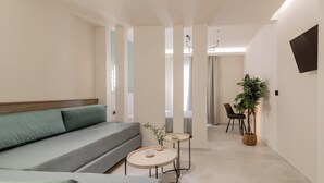 A modern, minimalist hotel room with a sofa, two small round tables, a potted plant, and a TV mounted on the wall. - Hera Zakynthos Hotel (Zakynthos)