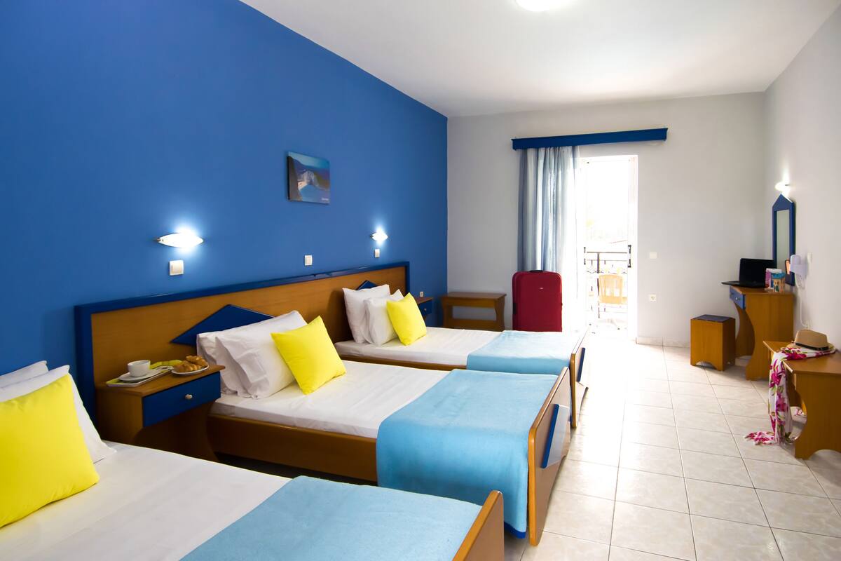 A hotel room with three beds, a wooden headboard, a nightstand with a phone, a red chair, a desk with a computer, and a window with curtains.