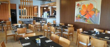 A restaurant with multiple tables and chairs, a bar area, and a large floral painting on the wall.