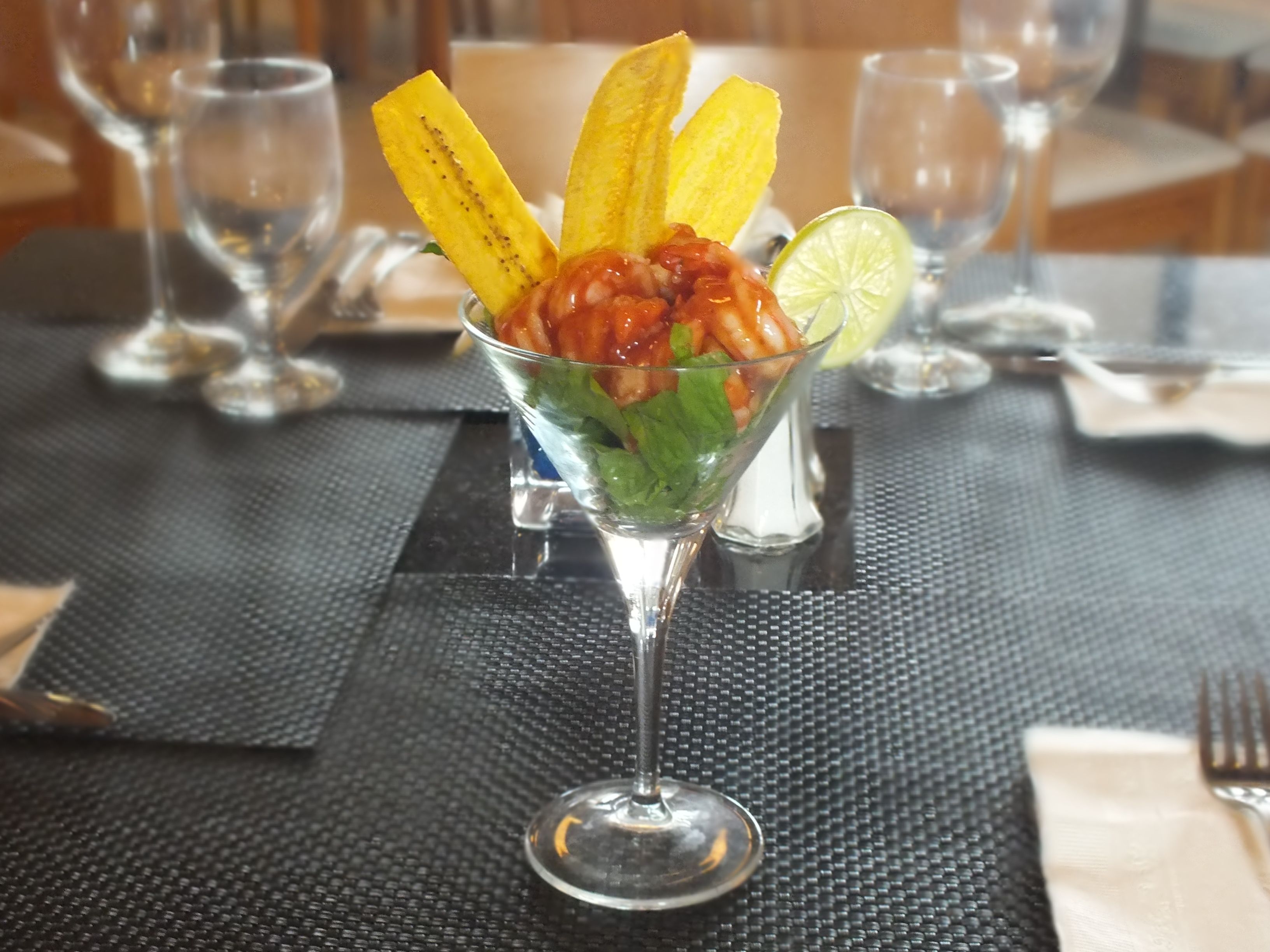 Un verre à martini, décoré d’un fruit jaune, peut-être une mangue, et d’une tranche de citron vert, posé sur une table recouverte d’une nappe noire.